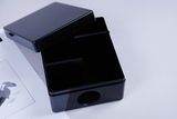 Das Bild zeigt eine schwarze, rechteckige Box mit einem abnehmbaren Deckel und zwei Trennwänden im Inneren. Neben der Box liegt ein Blatt Papier, das eine schwarz-weiße Abbildung einer Pflanze zeigt.