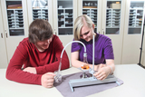Das Bild zeigt zwei Jugendliche, die gemeinsam an einem Experiment arbeiten. Sie verwenden ein chemisches Apparate-Set, wobei der Junge eine Flüssigkeit in ein Becherglas hält und das Mädchen am Experimentiertisch arbeitet.