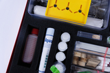 Das Bild zeigt ein Experimentier-Kit mit verschiedenen Materialien und Geräten, darunter Flaschen, Chemikalien und Behälter, die in einem gepolsterten Fach angeordnet sind. Es ist darauf ausgelegt, Schülern praktische Experimente und wissenschaftliche Entdeckungen zu ermöglichen.