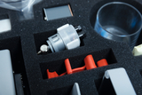 Das Bild zeigt eine Fächeraufteilung in einem Schaumstoffgehäuse, in dem verschiedene Experimentiermaterialien untergebracht sind. Zu den sichtbaren Objekten gehören ein silberner Hydraulikzylinder, rote Kunststoffteile und ein durchsichtiges Glas.