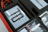 Das Bild zeigt eine Experimentierstation, die in einem Schaumstoff-Insert eines Koffers untergebracht ist. Zu sehen sind eine Platine mit Verbindungsstellen, weiße Komponenten und Kabel, die für wissenschaftliche Experimente im Bildungsbereich verwendet werden.