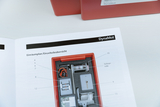 Das Bild zeigt eine Anleitung für ein Experimentiergerät namens DynaMot, das detaillierte Informationen zu den enthaltenen Einzelteilen bietet. Auf der Abbildung sind verschiedene Komponenten wie Motor und Schnurrolle in einem roten Gehäuse dargestellt.
