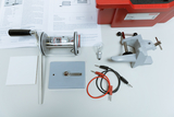 Das Bild zeigt eine Auswahl von Experimentiermaterialien, darunter einen Motor, eine Glühbirne, Kabel und eine Klemme auf einem Tisch. Zusätzlich sind ein Informationsblatt und eine aufbewahrende Box sichtbar, die die Materialien für Bildungszwecke zusammenfassen.