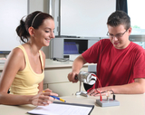 Das Bild zeigt zwei Schüler in einem Klassenraum, die an einem Experiment arbeiten. Der Junge betätigt ein Gerät, während das Mädchen Notizen macht und lächelt, was auf eine engagierte Lernatmosphäre hinweist.
