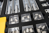 Das Bild zeigt eine sortierte Box mit Experimentiermaterialien, die für verschiedene physikalische Experimente genutzt werden können. In der Box sind Gewichte in transparenten Fächern sowie verschiedene Halterungen und Bauteile untergebracht.