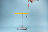 Das Bild zeigt eine experimentelle Vorrichtung zur Durchführung physikalischer Messungen. Eine Waage mit einem Hakengewicht hängt an einem gelben Balken, der auf einem stabilen Standfuß montiert ist, während ein Handgriff zur Justierung sichtbar ist.