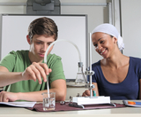 Auf dem Bild sind ein Schüler und eine Lehrerin zu sehen, die gemeinsam an einem Experiment arbeiten. Der Schüler fokussiert sich auf das Pipettieren einer Flüssigkeit in einen Becher, während die Lehrerin ihm lächelnd zuschaut.