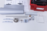Das Bild zeigt eine Sammlung von Experimentiermaterialien, die auf einem Tisch angeordnet sind, einschließlich eines roten Koffers, verschiedenen Metall- und Kunststoffteilen, einem Glasbehälter und einer Anleitung. Diese Materialien dienen vermutlich der Durchführung von Wärmelehre-Experimenten im Bildungsbereich.