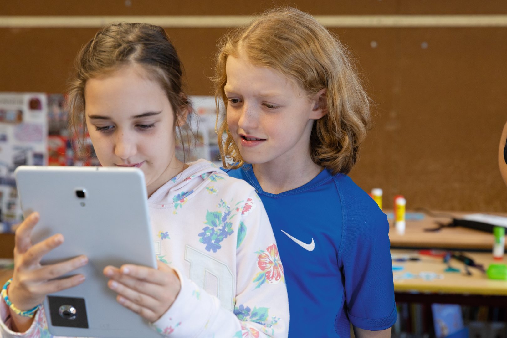 Auf dem Bild sind zwei Kinder zu sehen, die gemeinsam auf ein Tablet schauen. Die Atmosphäre wirkt neugierig und freundlich, im Hintergrund sind künstlerische Materialien und Poster zu erkennen, was auf eine kreative Lernumgebung hindeutet.