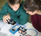 Das Bild zeigt zwei Schüler, die an einem Tisch experimentieren und ein Smartphone nutzen, um ein Experiment zu beobachten. Neben ihnen befinden sich Messgeräte und eine Konstruktion mit Spulen, die für eine technische oder physikalische Untersuchung verwendet wird.