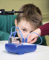 Das Bild zeigt einen Jungen, der konzentriert an einem Experimentiergerät arbeitet, während er von einer anderen Person unterstützt wird. Das Gerät ist mit bunten Kabeln verbunden, die für wissenschaftliche Experimente verwendet werden.