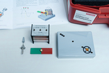 Das Bild zeigt verschiedene Experimentiermaterialien zur Demonstration von Induktion und Wechselspannung, darunter eine Spule, einen Magneten und ein Steuergerät. Neben diesen Materialien liegt ein Handbuch auf, das erläuternde Diagramme und Informationen enthält.