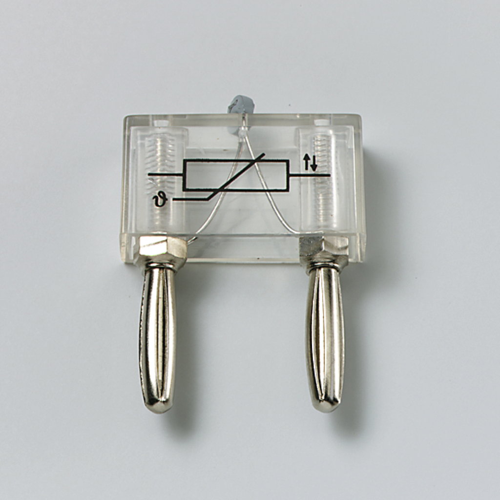 Das Bild zeigt ein transparentes Gerät mit zwei silbernen Anschlüssen und einem einfachen Schaltkreis in der Mitte. Es ist wahrscheinlich ein Experimentiergerät zur Demonstration von elektrischen oder physikalischen Prinzipien im Bildungsbereich.