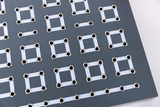Das Bild zeigt eine flache, graue Platte mit mehreren Anordnung von silbernen Kontaktlöchern und weißen, rechteckigen Leiterbahnen. Diese wird vermutlich für experimentelle Elektroanwendungen im Bildungsbereich verwendet.