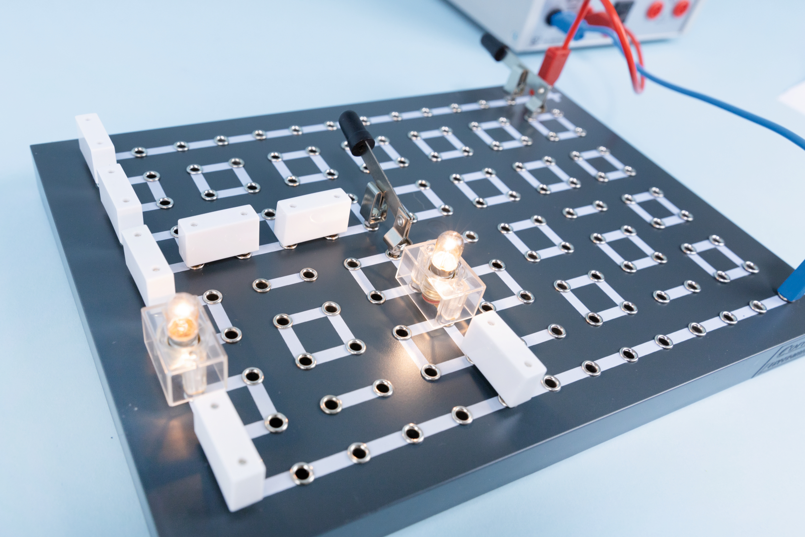 Das Bild zeigt ein Experimentierbrett mit verschiedenen elektrischen Verbindungen, auf dem einige kleine Glühbirnen leuchten. Es sind mehrere Kunststoffkomponenten und Klemmen angebracht, die für das Testen und Verstehen elektrischer Schaltungen verwendet werden.