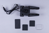 Das Bild zeigt eine schwarze Halterung mit einem Kabel und mehreren Platten, die vermutlich für Experimente im Bildungsbereich genutzt werden. Die Halterung hat eine klammerartige Form, während die Platten aus unterschiedlichen Materialien bestehen.