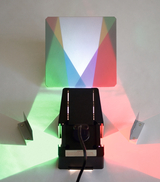 Das Bild zeigt ein Experimentiergerät, das aus einer Lichtquelle und einer prismatischen Fläche besteht. Es erzeugt farbige Lichtstrahlen in Rot, Grün und Blau, die an die Wand projiziert werden, um Lichtbrechung und -mischarbeiten zu demonstrieren.