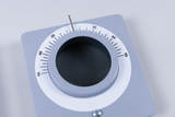 Das Bild zeigt ein grau lackiertes Messinstrument mit einer runden Skala, auf der Winkel von 0 bis 90 Grad angezeigt werden. In der Mitte befindet sich eine Öffnung, die möglicherweise zur Durchführung von Experimenten oder Messungen dient.
