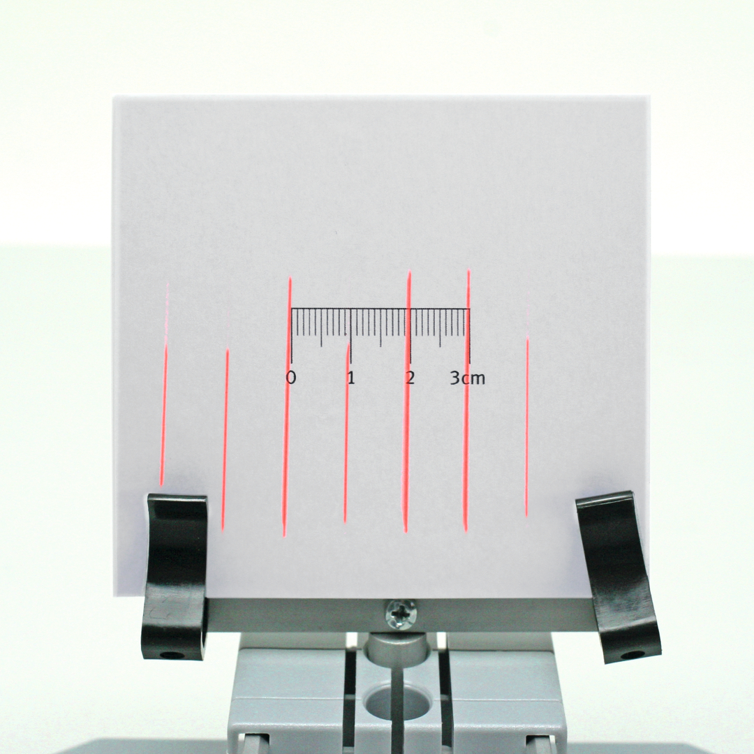 Das Bild zeigt ein weißes Blatt mit roten, parallelen Linien und einer Maßstabsmarkierung von 0 bis 3 cm. Es ist auf einer Halterung befestigt, die für Experimente im Bildungsbereich genutzt wird.