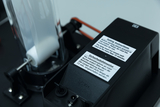 Das Bild zeigt ein technisches Gerät mit einer transparenten Kammer und einem elektronischen Bedienfeld, das Sicherheitsanweisungen in Englisch und Deutsch enthält. Es scheint für experimentelle Zwecke im Bildungswesen konzipiert zu sein und hat elektrische Anschlüsse.