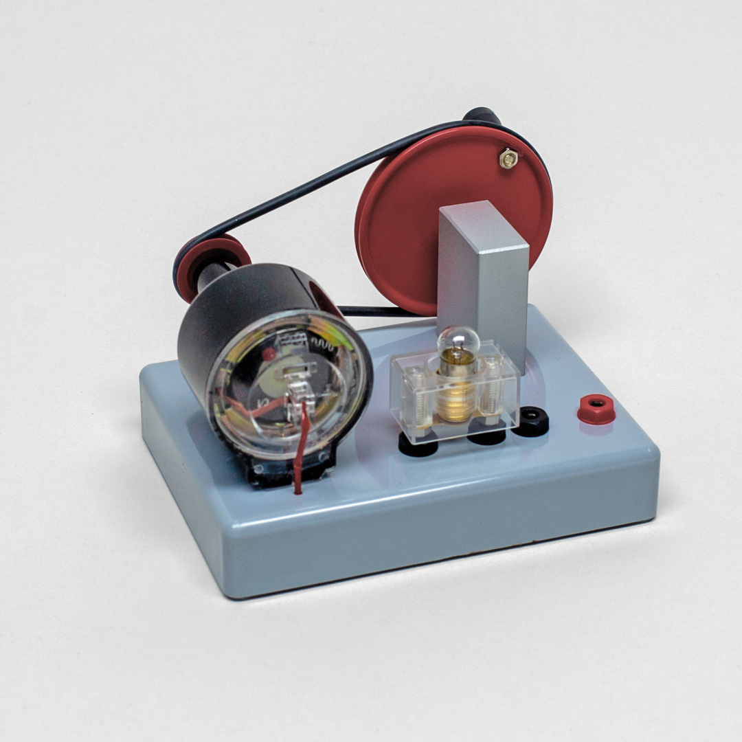 Das Bild zeigt ein Experimentiergerät, das vermutlich zur Demonstration physikalischer Prinzipien wie Elektromagnetismus oder Mechanik dient. Es besteht aus einer Basisplatte mit verschiedenen Komponenten, darunter ein Motor, ein Pulley-System und Anschlüsse für elektrische Verbindungen.