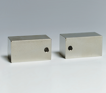Das Bild zeigt zwei rechteckige Metallblöcke, die eine polierte Oberfläche haben und jeweils ein Loch auf einer der Seiten aufweisen. Diese Blöcke könnten für physikalische Experimente oder für den Einsatz in der Bildungsforschung verwendet werden.