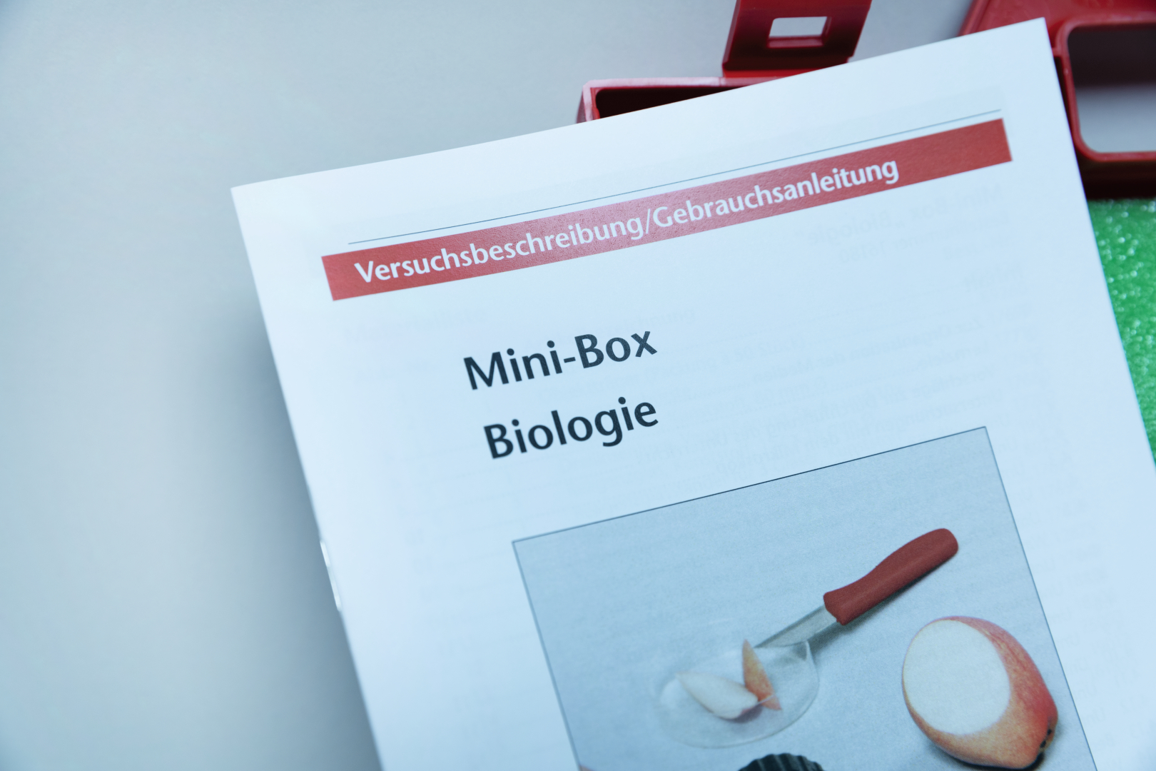 Das Bild zeigt die Gebrauchsanleitung einer Mini-Box für biologischen Experimentierbedarf im deutschen Bildungswesen. Auf der Anleitung ist ein Bild zu sehen, das eine Essenzubereitung mit einem Messer und einem Apfel zeigt.