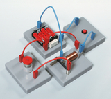 Das Bild zeigt ein Experimentiergerät, das aus mehreren grauen Basiselementen besteht, die miteinander verbunden sind. Auf den Platten befinden sich eine Batterieeinheit, ein Schalter und eine Kupferspule, die für elektrische Experimente im Bildungswesen verwendet werden.