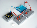 Das Bild zeigt ein Experimentier-Setup mit einer elektrochemischen Zelle, bestehend aus einer Wasserschicht mit blauer Flüssigkeit und verschiedenen Anschlüssen. Es sind Batterien, Verbindungsdrähte und Messinstrumente zur Durchführung von Experimenten sichtbar.