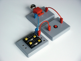 Das Bild zeigt ein Experimentier-Set, das aus mehreren grauen Platten, Kabeln, einer metallischen Kugel und einer Schaltfläche besteht. Es dient der Veranschaulichung elektrischer Schaltungen und kann im Bildungsbereich für praktische Übungen eingesetzt werden.
