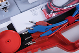 Das Bild zeigt eine Anordnung von elektrischen Experimentierkomponenten, darunter bunte Kabel mit Stecker, die für den Einsatz in Bildungsexperimenten gedacht sind. Ein roter Halter und eine Verpackung sind ebenfalls im Bild zu sehen, was auf eine strukturierte Präsentation der Materialien hinweist.