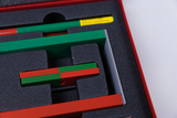 Das Bild zeigt ein Set aus verschiedenen geometrischen Körpern in Grün und Rot, die auf einem grauen Schaumstoffpolster angeordnet sind. Die Farben und Formen sind vermutlich für experimentelle Zwecke im Bildungsbereich konzipiert.