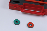 Das Bild zeigt eine rote Box mit Schaumstoffeinlage, die magnetische Scheiben in verschiedenen Farben enthält. Zwei dieser Scheiben, eine grün und eine rot, liegen auf einem weißen Untergrund vor der Box.