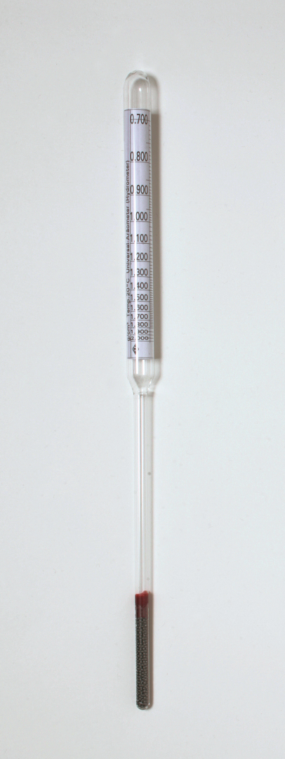 Das Bild zeigt ein Hydrometer, ein transparentes Messgerät mit einer Graduierung zur Bestimmung der Dichte von Flüssigkeiten. Am unteren Ende enthält es eine kleine Menge einer dunklen Flüssigkeit, die als Referenz dient.