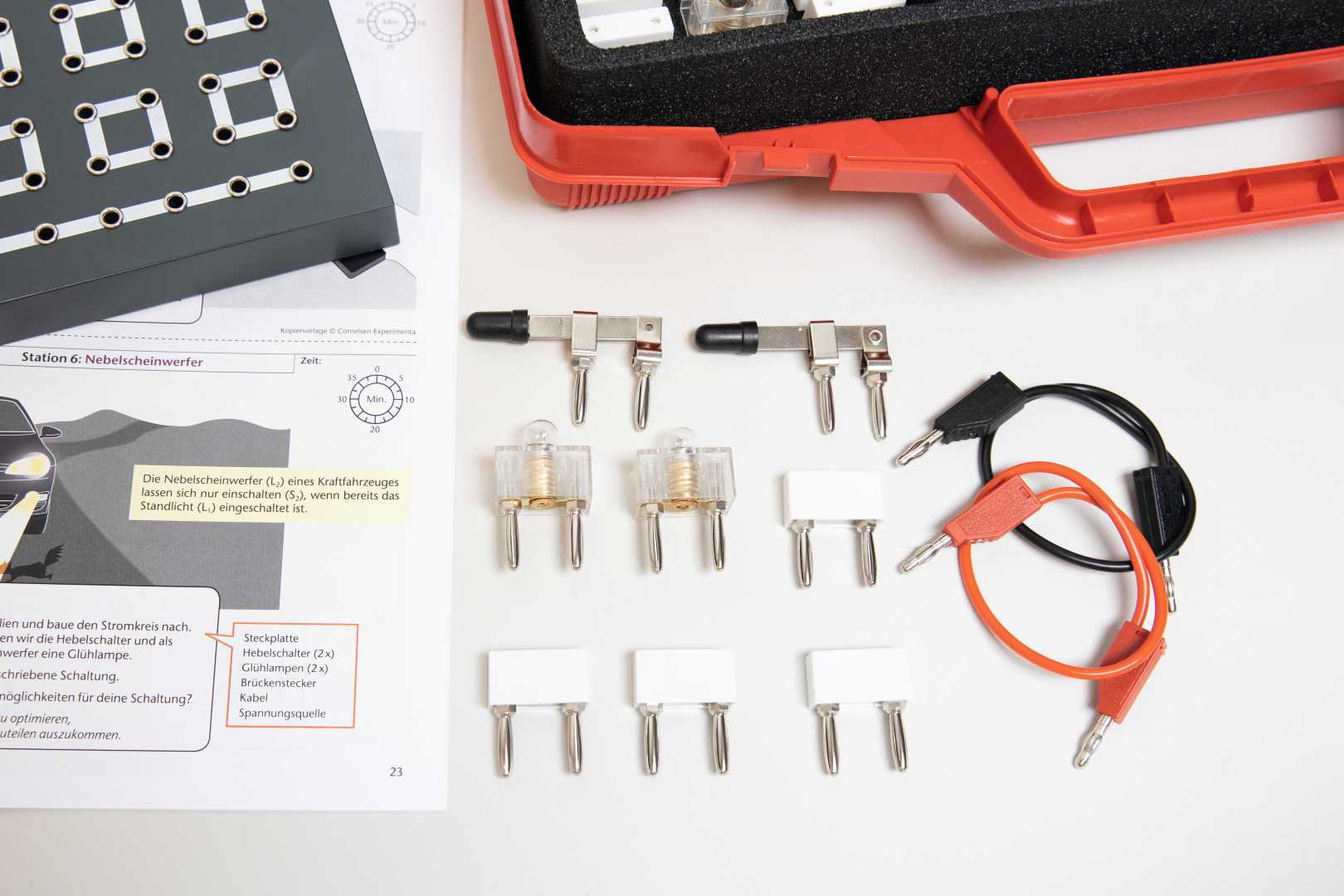 Das Bild zeigt verschiedene elektronische Komponenten, darunter Schalter, Glühlampen und Verbindungskabel, die für Experimente im Bildungsbereich verwendet werden. Neben den Materialien liegt eine Anleitung mit Informationen zur Durchführung eines Experiments zur Steuerung von Nebelscheinwerfern.