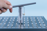 Das Bild zeigt eine Hand, die einen Metallstab über einem silbernen Zylinder hält, der auf einer Platte mit elektrischen Kontakten steht. An dem Zylinder hängt ein kleiner Körper, was auf ein Experiment zur Demonstration von physikalischen Prinzipien hinweist.