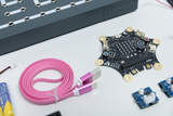 Das Bild zeigt verschiedene elektronische Komponenten, darunter ein Calliope Mini Board, ein pinkes USB-Kabel und mehrere Sensoren. Diese Materialien sind für Experimente im Bildungsbereich konzipiert und fördern das Lernen in Informatik und Technik.