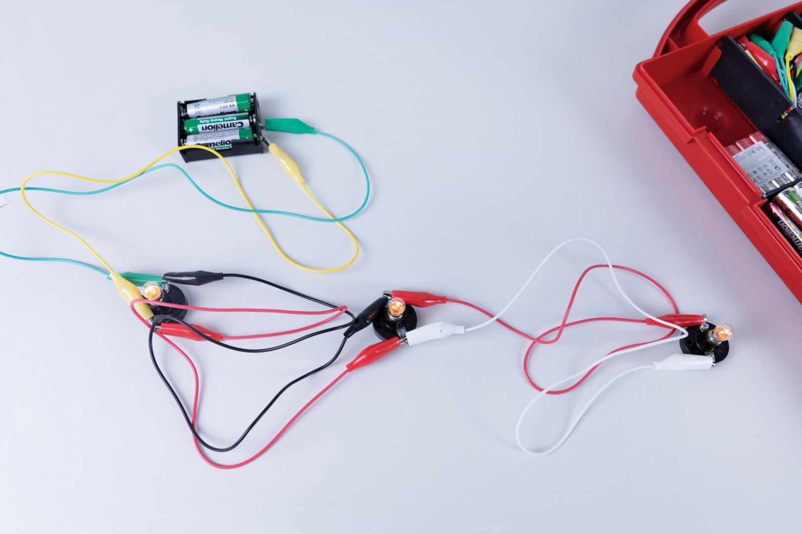 Das Bild zeigt eine Anordnung von elektrischen Bauteilen, einschließlich einer Batteriehalterung mit Batterien und mehreren Kabeln, die über Krokodilklemmen verbunden sind. Ein Teil der Schaltung scheint eine Lampe zu betreiben, die durch die Verbindungen aktiviert wird.