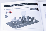 Das Bild zeigt ein Experimentier-Setup zur Bestimmung des Humusgehalts in Bodenproben. Es enthält verschiedene Laborgeschirre und Sicherheitshinweise, darunter Warnsymbole für Gefahren und das Tragen einer Schutzbrille.