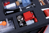 Das Bild zeigt ein Set mit verschiedenen Experimentiermaterialien, das in einem Schaumstoffpolster angeordnet ist. Zu sehen sind unter anderem ein Glaskolben, ein Metallglockenläuten, eine orangefarbene Knopfsonde und diverse kleine Werkzeuge.