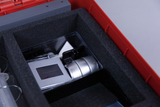 Das Bild zeigt eine rote Box, die mit schwarzem Schaumstoff gepolstert ist und verschiedene Experimentiergeräte enthält. Sichtbar sind unter anderem ein Gerät mit Display und eine silberne Zylindereinheit, die für Bildungs-Experimente verwendet werden können.