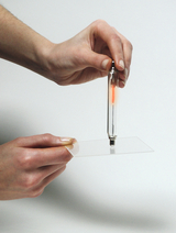 Das Bild zeigt eine Hand, die ein Röhrchen mit einer roten Substanz hält, während sie es über eine transparente Oberfläche positioniert. Das Experiment scheint darauf abzuzielen, eine Chemie- oder Physikreaktion zu demonstrieren.