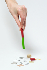 Das Bild zeigt eine Hand, die einen mehrfarbigen Magnetstab über einer Anordnung von verschiedenen Materialien hält, darunter Metallstückchen, Holz und Papier. Die Darstellung ist ideal für Experimente im Bildungsbereich, insbesondere zur Veranschaulichung von Magnetismus.