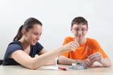 Das Bild zeigt zwei Schüler, die gemeinsam an einem Experimentiergerät arbeiten. Der Junge hält eine Schnur, während das Mädchen aufmerksam zuschaut und eine Büroklammer positioniert, was auf eine praktische Lernaktivität hinweist.