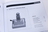 Das Bild zeigt die Komponenten eines Versuchsaufbaus zur Funktion einer elektrischen Klingel, inklusive Schalter und Klingelmechanismus. Daneben sind die benötigten Materialien und zusätzliche Anforderungen aufgelistet.