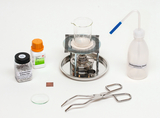 Das Bild zeigt ein Set aus verschiedenen Laborgeräten und Chemikalien, das für Experimente im Bildungsbereich verwendet wird. Zu den Gegenständen gehören ein Becherglas, ein Bunsenbrenner, Siedesteinchen, eine Pipette, entmineralisiertes Wasser und eine Zange.