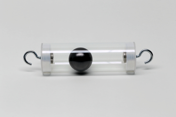 Das Bild zeigt einen transparenten Zylinder mit einer schwarzen Kugel in der Mitte, die durch zwei Metallhaken auf beiden Seiten des Zylinders gehalten wird. Dieses Experimentiergerät könnte zur Veranschaulichung physikalischer Prinzipien wie Schwingungen oder Gleichgewicht verwendet werden.