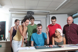 Das Bild zeigt eine Gruppe von sechs jungen Menschen und einem Erwachsenen, die einen experimentellen Aufbau an einem Tisch betrachten. Der junge Mann in der Mitte erklärt etwas, während die anderen interessiert zuhören oder mit Geräten interagieren.