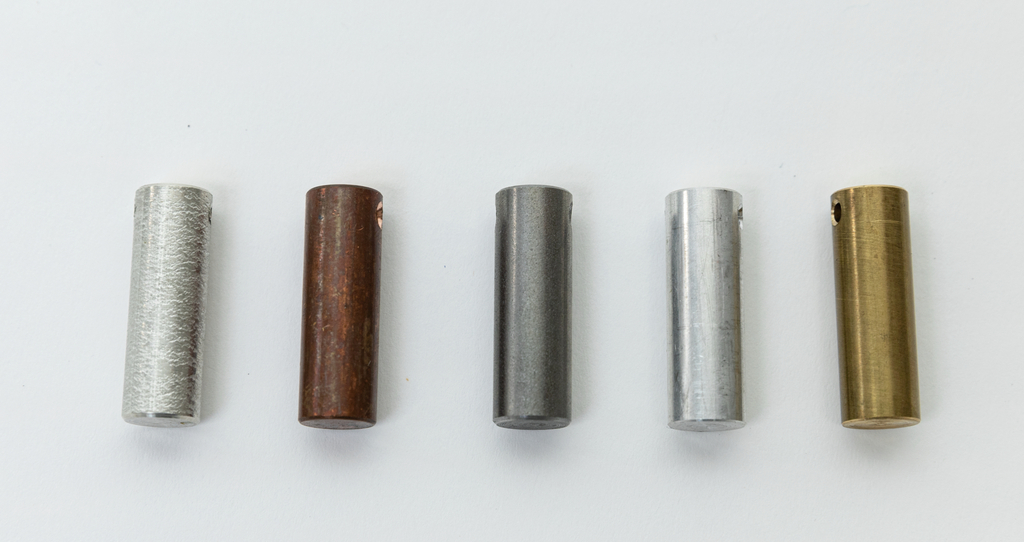 Das Bild zeigt fünf unterschiedlich gefärbte Zylinder aus Metall, die in einer Reihe angeordnet sind. Sie variieren in Farbe und Material und könnten für experimentelle Zwecke im Bildungswesen verwendet werden.