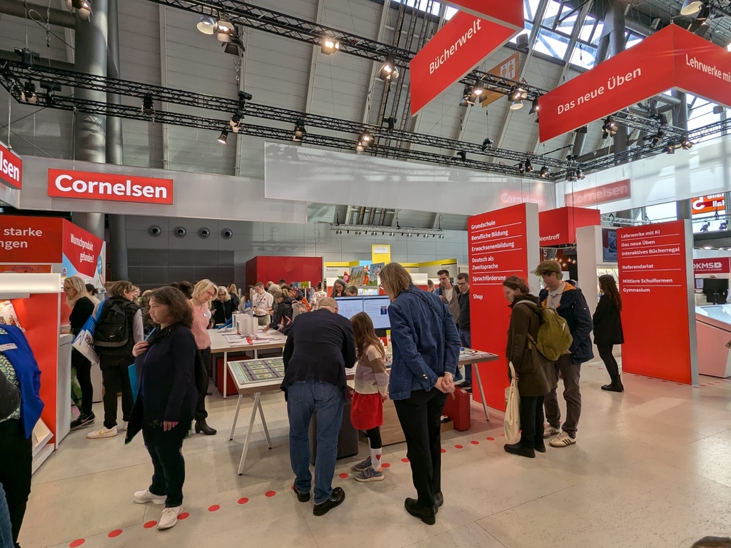 Das Bild zeigt einen belebten Stand von Cornelsen auf einer Bildungsmesse, wo Besucher verschiedene Lernmaterialien und Angebote erkunden. Die Wandtafeln informieren über Produkte für Grundschule, berufliche Bildung und Sprachförderung.